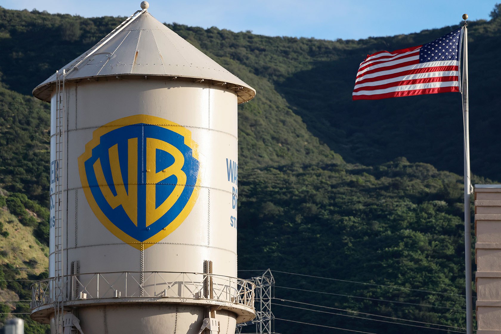 The yellow and blue WB logo on a beige water tower; an American flag is positioned beside but behind the tower.