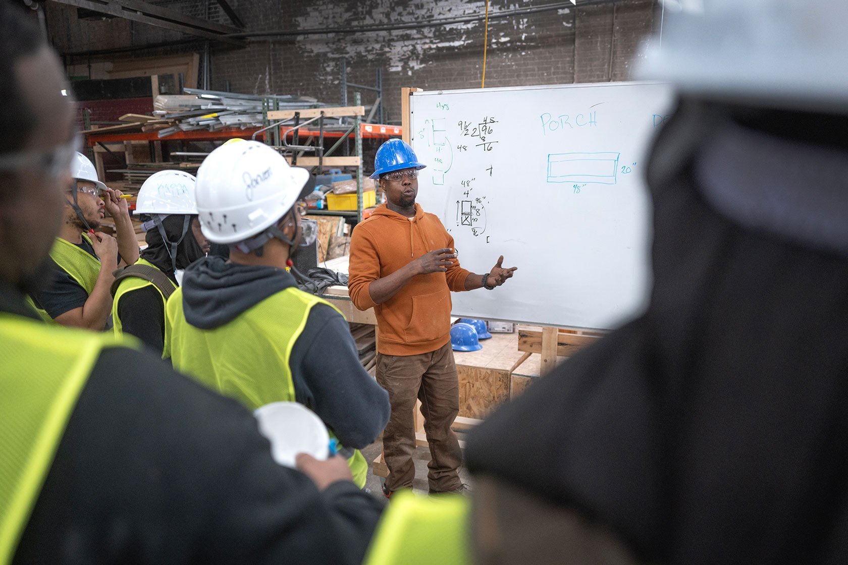 An instructor standing in front of a group of students dressed for construction work. The whiteboard behind him features instructions for building a porch.